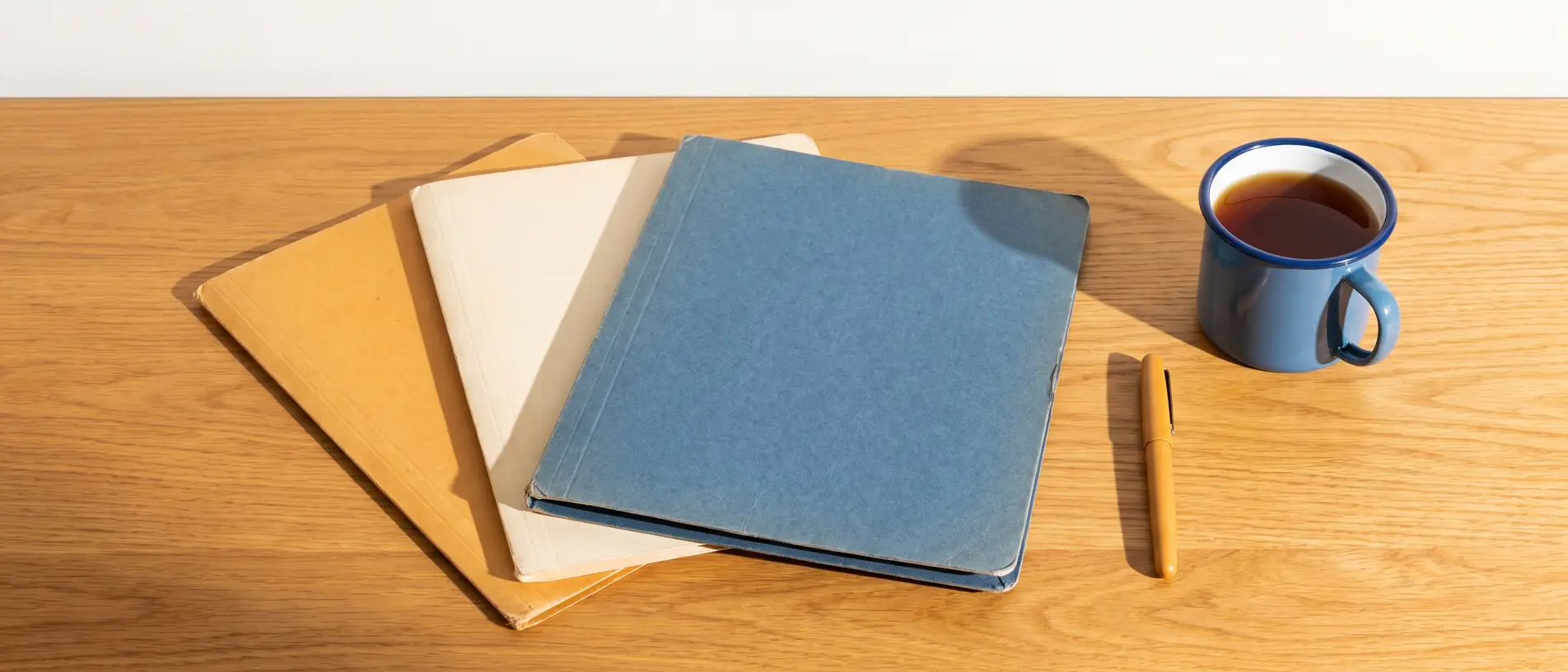 Three blank card document folders of increasing thickness fanned across a sunlit wooden table in ochre, cream and denim blue, with a pen and a blue enamel mug beside them.