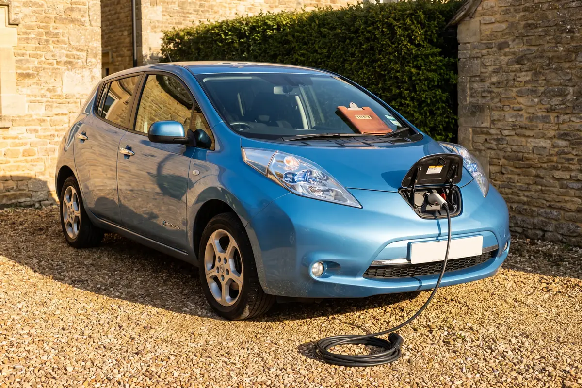 A second-hand blue electric hatchback parked on a bright gravel driveway in hard morning sunlight, its charging flap open at the front with the charging cable running down to a loose coil on the gravel, a honey-coloured stone wall and clipped green hedge behind it, the kind of business car that sits in the main pool and is now written down at 14% a year.
